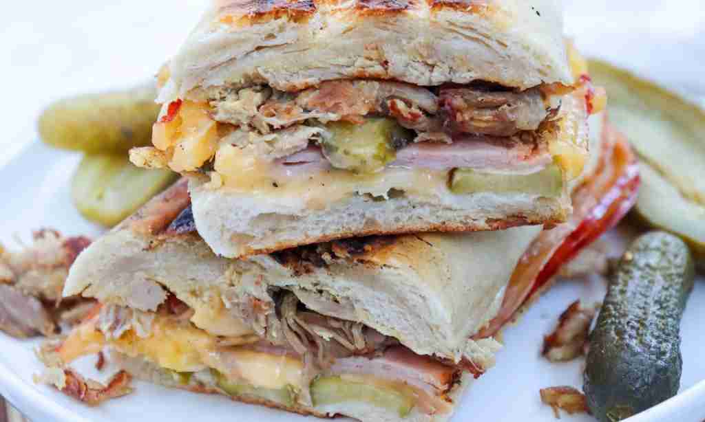 Cuban Sandwich Recipe A Cubana Sandwich recipe cute in half on a white plate with pickles